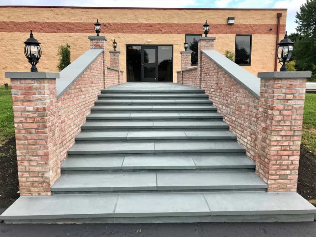 Wide bluestone stairs lead to a building entrance, bordered by red brick walls with black lantern fixtures on the pillars.