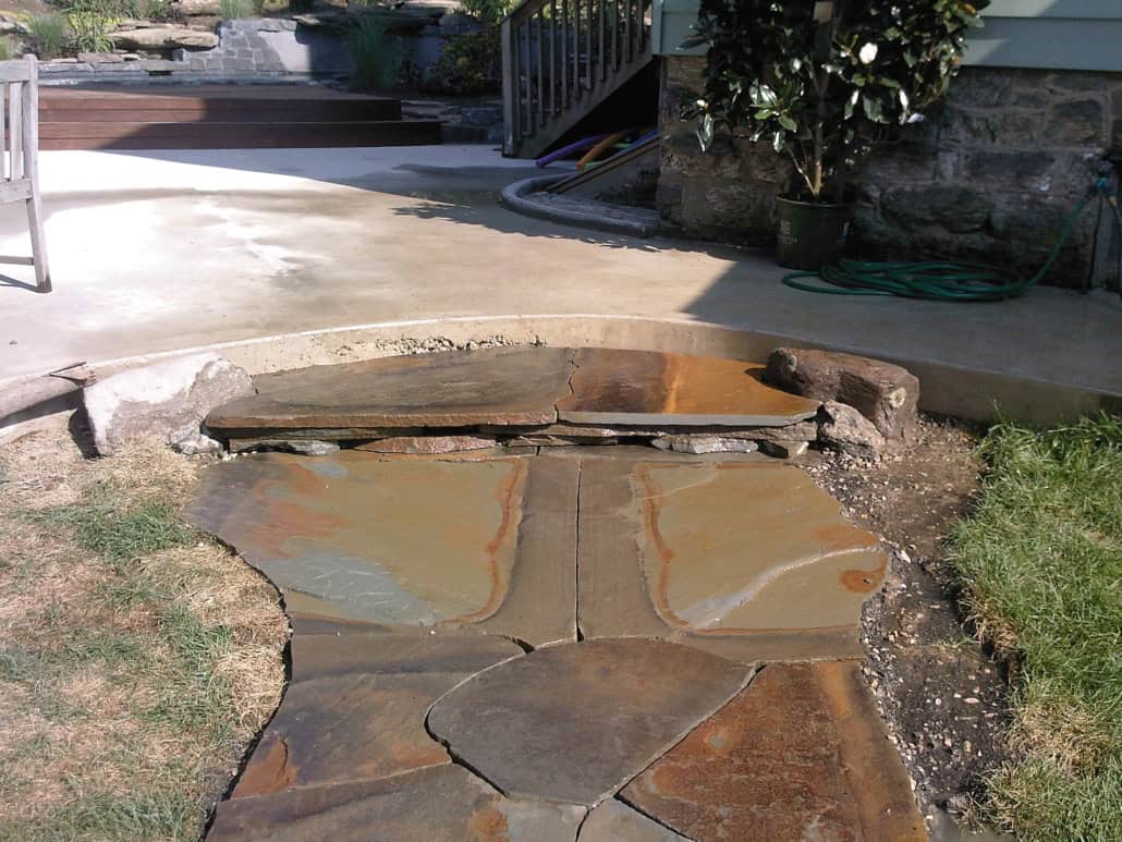 A path made of large, irregular flagstones in earthy brown and tan tones, leading to a curved concrete patio in a residential backyard.