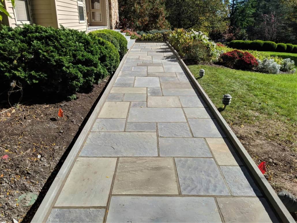 A custom-installed natural cleft bluestone walkway featuring a geometric pattern of rectangular pavers leading to a house entrance, bordered by neat garden beds and green shrubs.