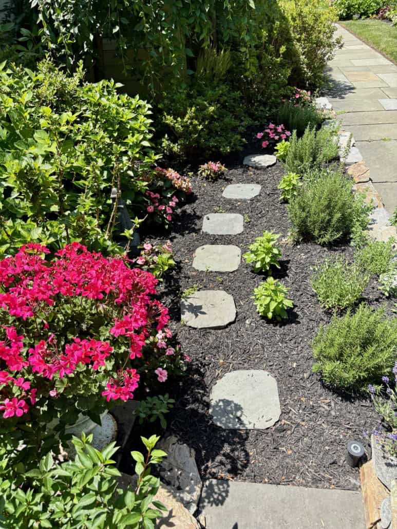 A winding garden path made of irregular natural stone stepping stones set in dark mulch, bordered by vibrant pink flowers and lush green landscaping shrubs.