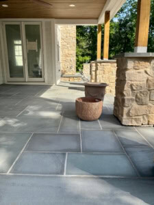 Modern covered patio featuring a thermal bluestone paver floor, natural stone pillars with wood posts, and a wood-paneled ceiling.