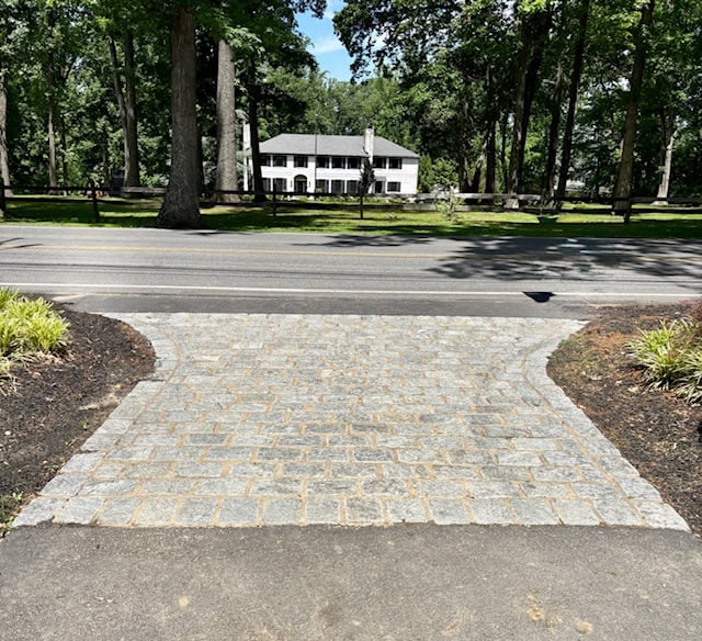 A newly installed Belgian block driveway apron with gray granite cobblestones laid in a classic running bond pattern, flaring out to meet a paved public road.