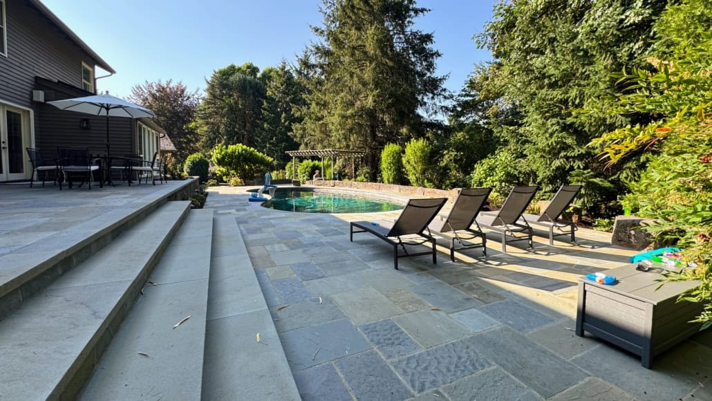 Multi-level bluestone patio with wide stone steps, lounge chairs, and an inground swimming pool surrounded by trees.